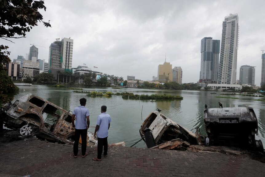 Two men stand beside lake with burnt out cars on shore.