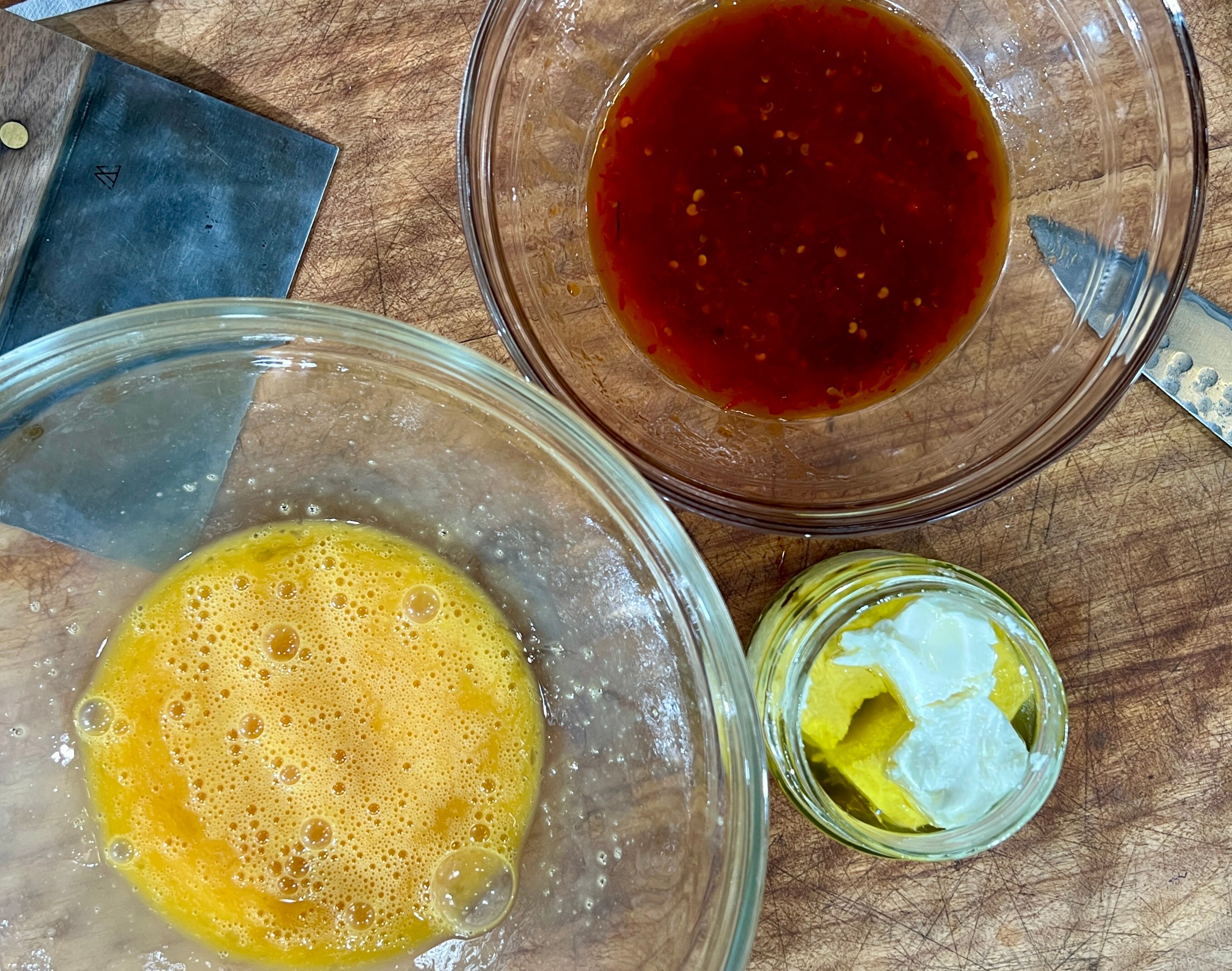A picture of chilli oil in a mixing bowl alongside beaten eggs in a mixing bowl and cheese in a jar on bench top.