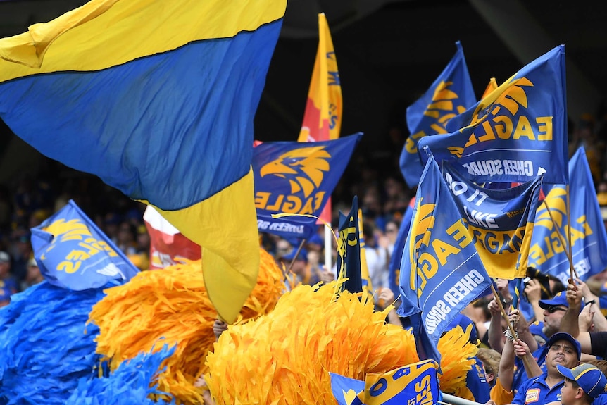 West Coast Eagles fans celebrate in the stands waving blue and yellow flags and pom poms.