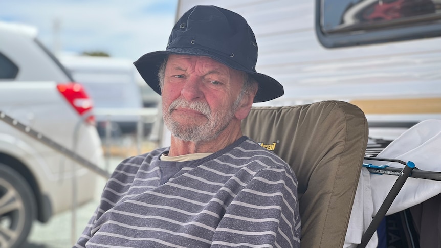 An older man wearing a black hat and stripy jumper sitting in front of a caravan.