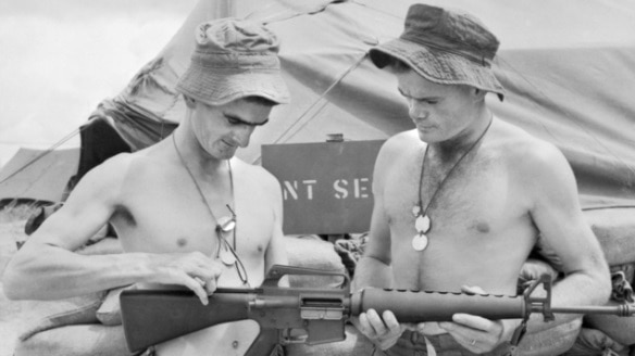 Two shirtless men look at a rifle in black and white photo