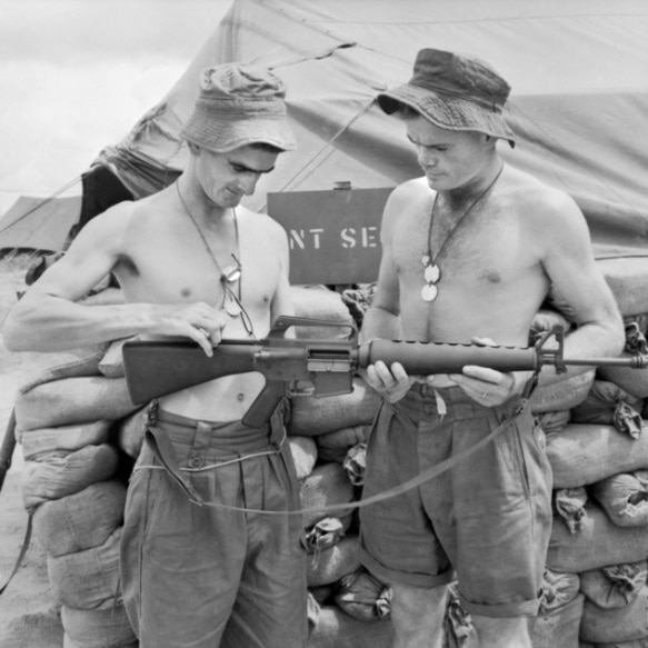 Two shirtless men look at a rifle in black and white photo
