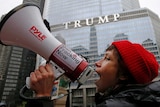 A woman yells on a megaphone and wears a red beanie, a trump property is visible in the background