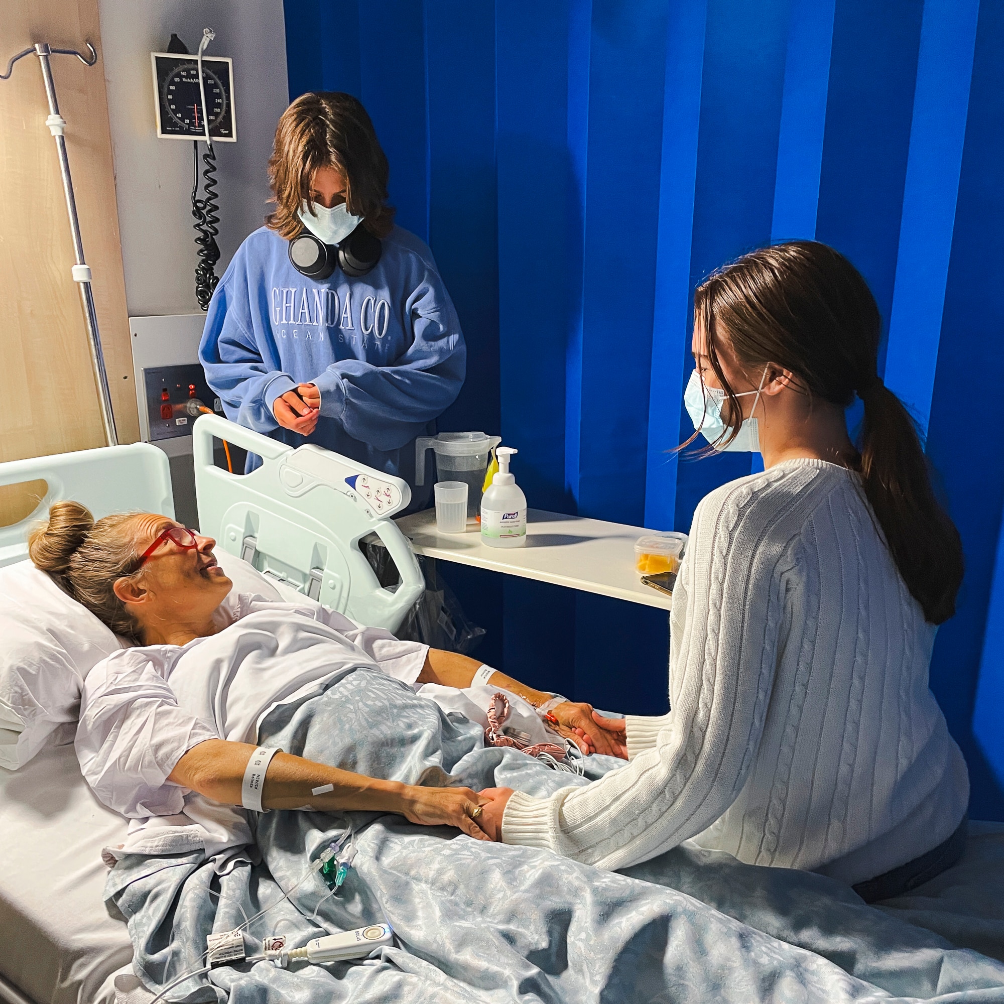 A woman lies in a hospital bed and two teenagers stand and sit by her bed. They're wearing masks.