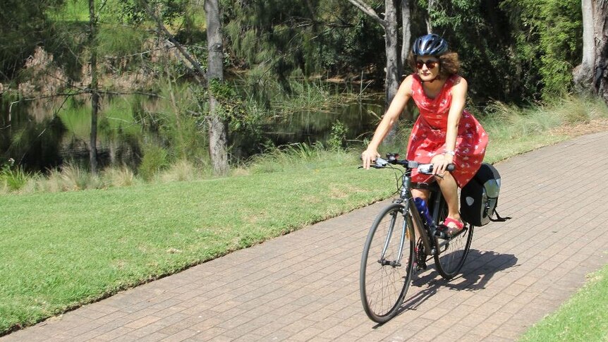A woman in a red dress rides a bicycle on a path lined with grass and trees.