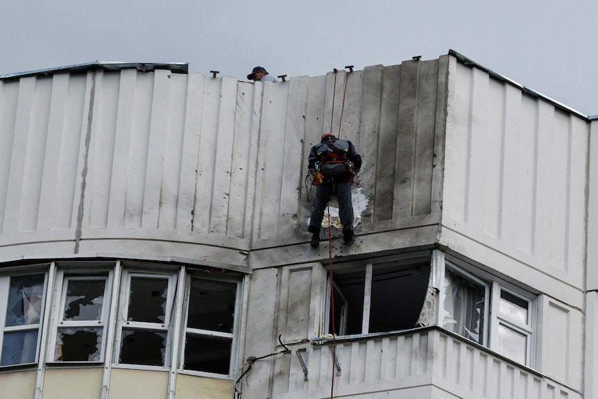 A worker abseils against a building to inspect drone damage. 
