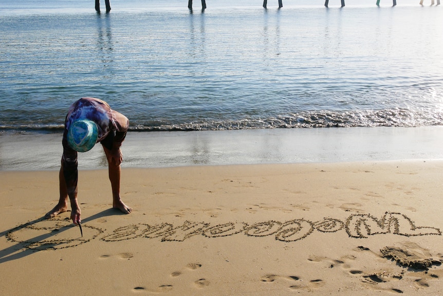A man uses a stick to write 'welcome' in the sand on the water's edge.