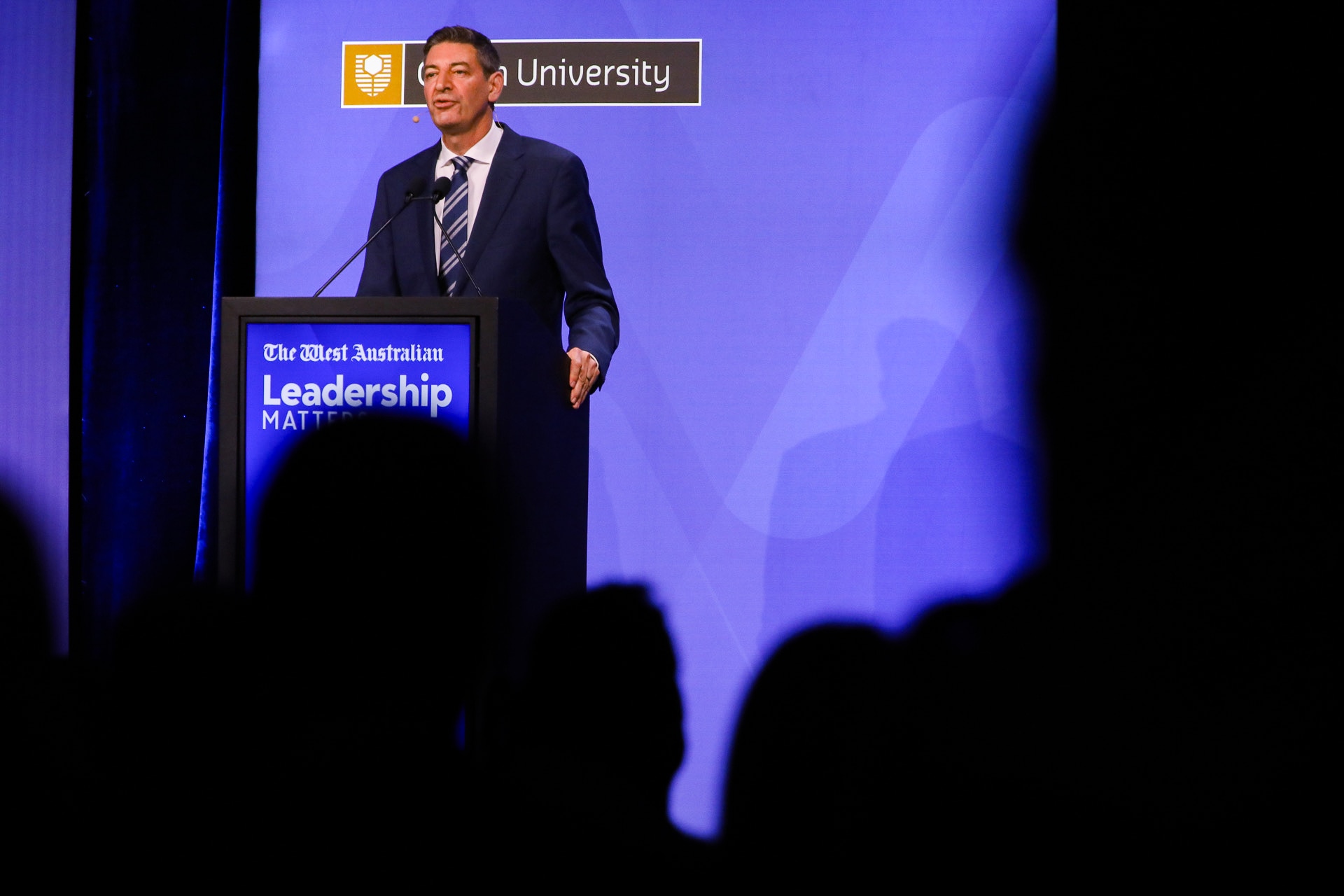 WA Opposition Leader Basil Zempilas speaks on stage at a podium with a blue background and shadows in the foreground.