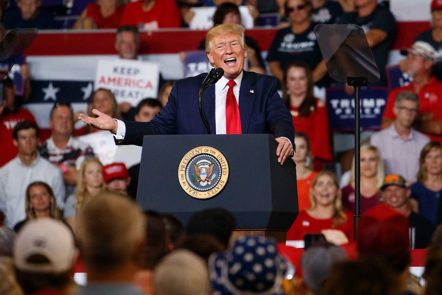 Donald Trump gestures with his right hand while speaking at a lectern. A crowd of people watch on.