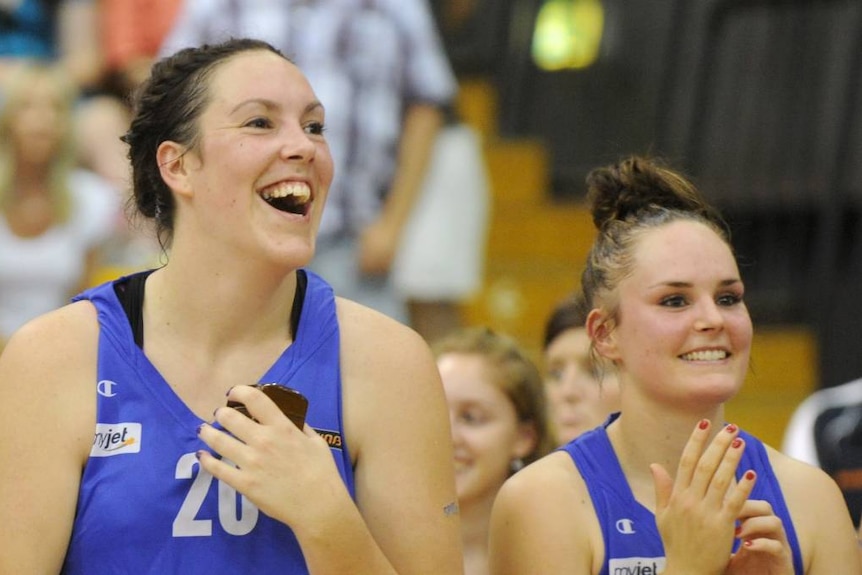 Two girls on the sidelines of a basketball court, in blue uniforms smiling and cheering.