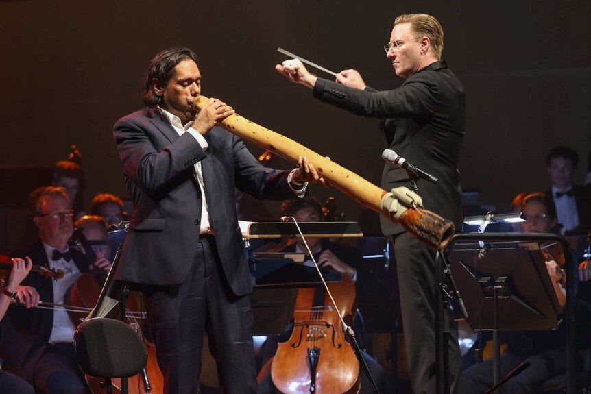 A man plays a didgeridoo with an orchestra.