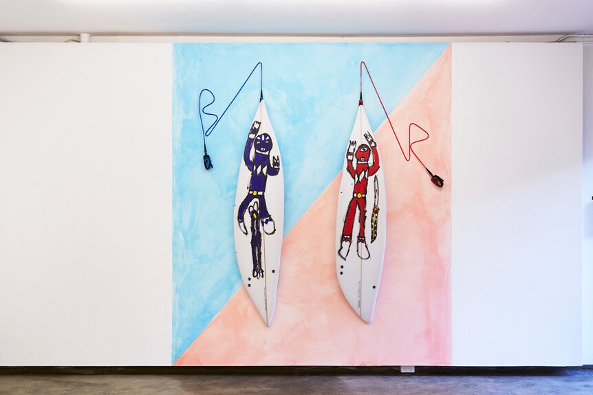 Two surfboards, emblazoned with paintings of the blue and red power rangers respectively, hang in a white gallery space.