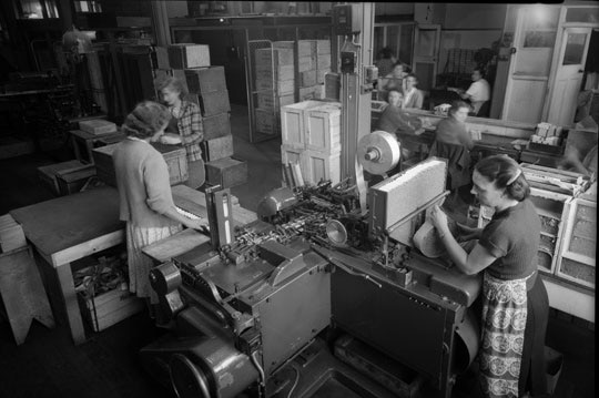 Black and white photo of women working at cigarette rolling machines inside a factory.