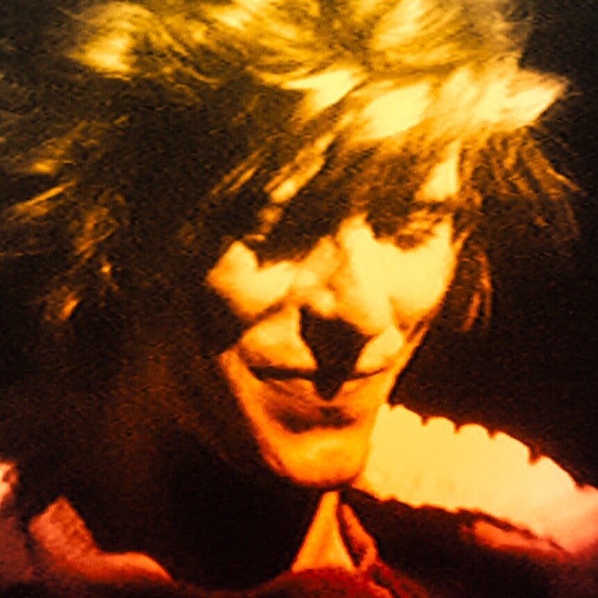 A close up of David Bowie with short tousled blonde hair smiling with eyes downcast.