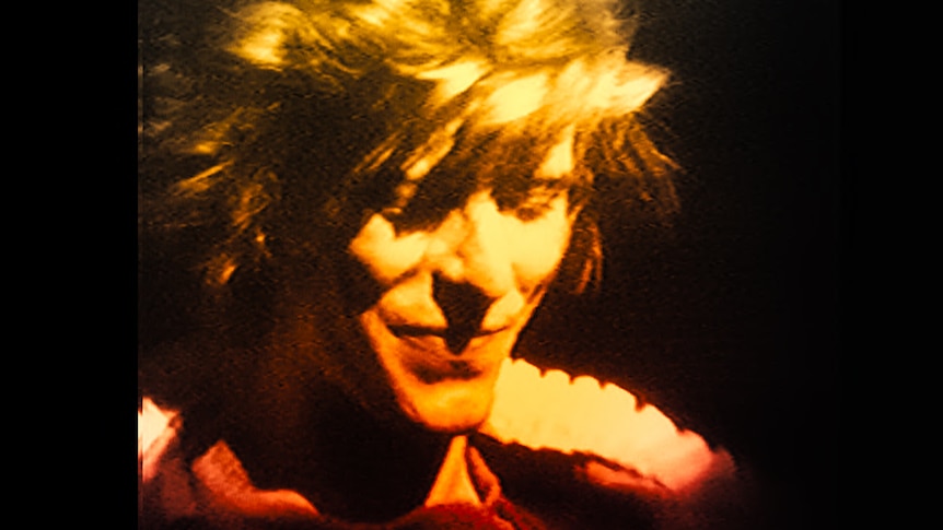 A close up of David Bowie with short tousled blonde hair smiling with eyes downcast.