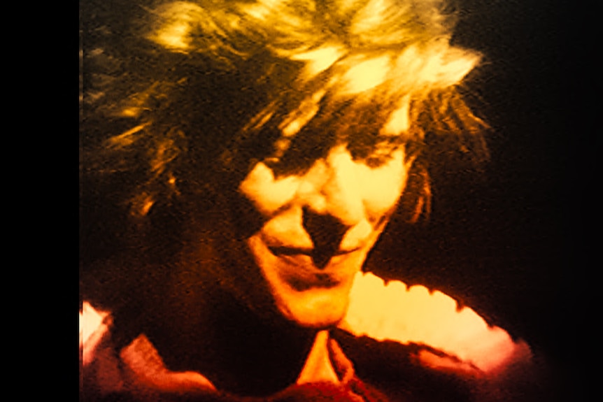 A close up of David Bowie with short tousled blonde hair smiling with eyes downcast.