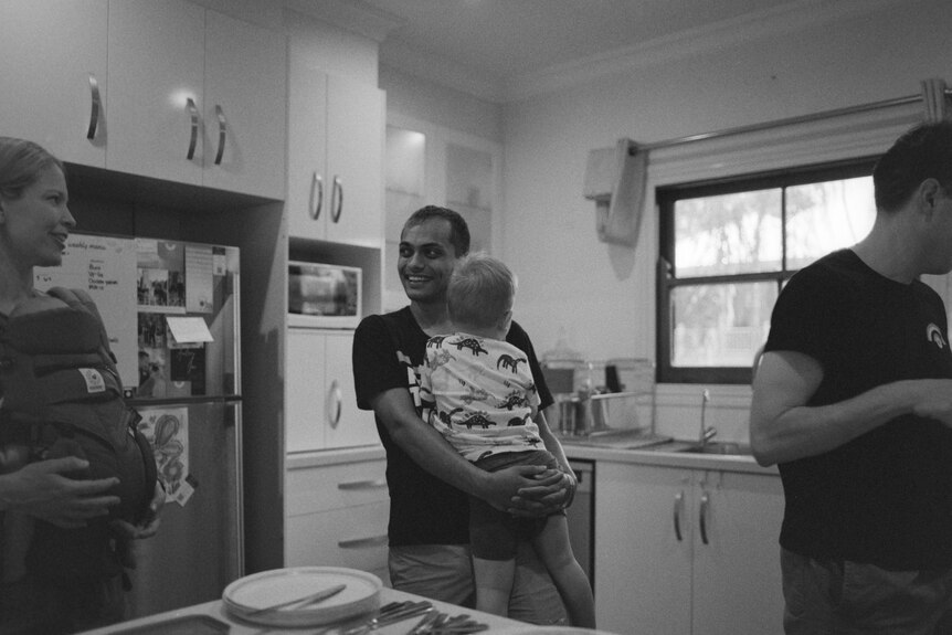 A man holds a toddler around a kitchen table. 