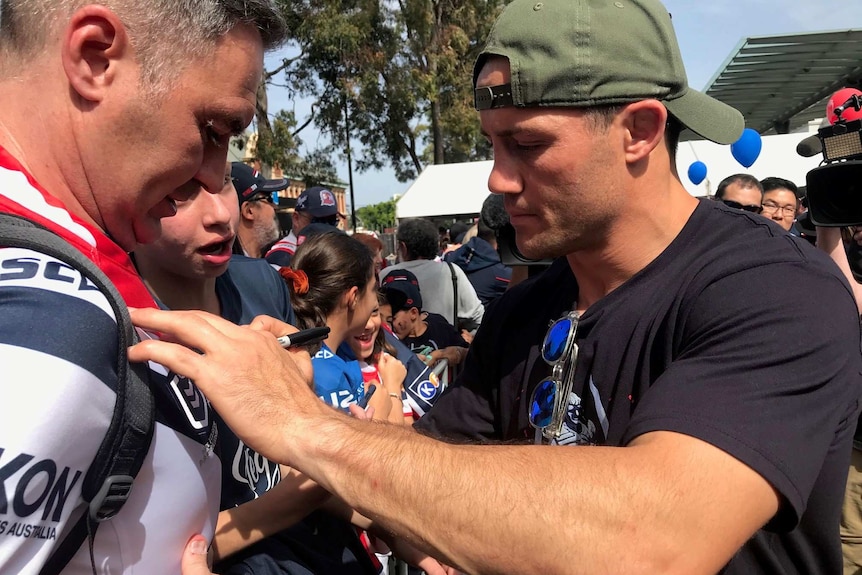 A man with his hat on backwards signs a fan's jersey.