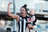 A woman wearing a black and white footy jumper who's carrying a child, raises her arm in celebration.
