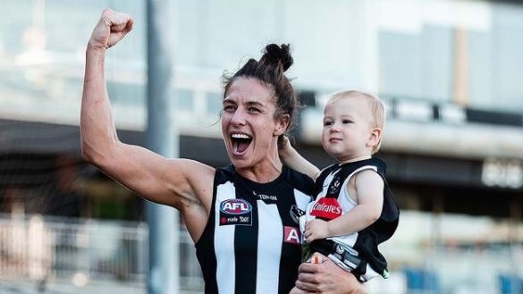 A woman wearing a black and white footy jumper who's carrying a child, raises her arm in celebration.