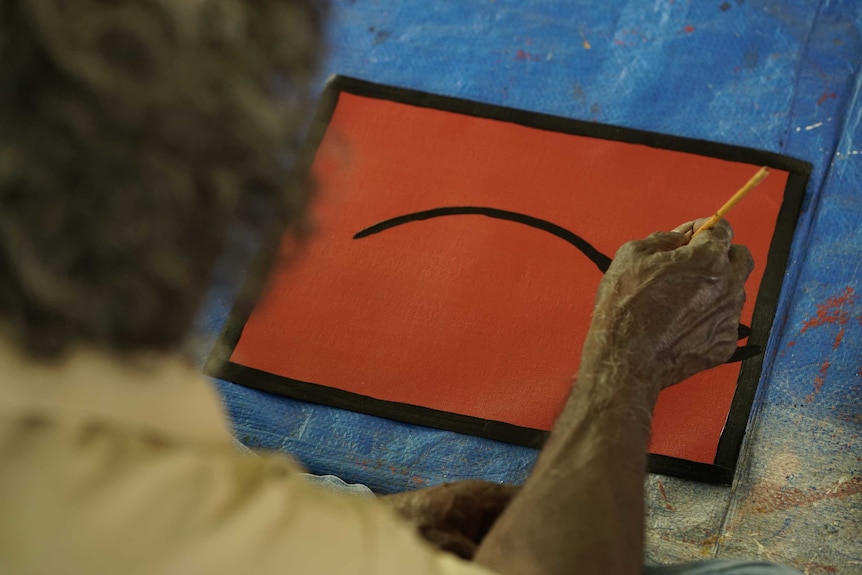 View from the back of an Indigenous man who is making long brush strokes with black paint on a red canvas