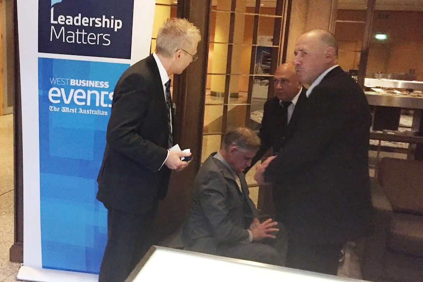 A man sits in a chair in a grey suit with three security officials in black suits standing around him.