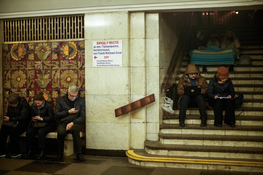 A group of men and women in winter clothing sit on benches and stairs in a tiled subway station, all looking at their phones.