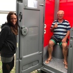 Volunteers celebrate arrival of a portable toilet by posing for happy photo one perched on top of the toilet with thumbs up.