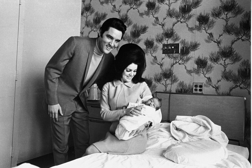 Priscilla holds Lisa Marie in her arms sitting on a bed as Elvis stands for the photo. 