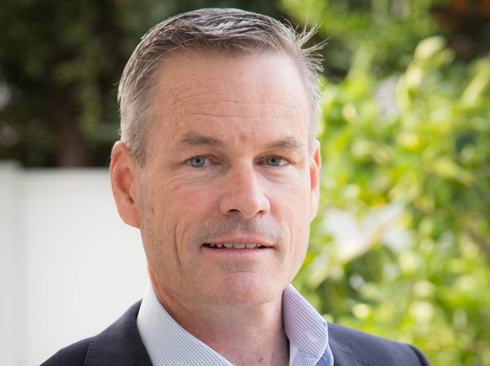 A head and shoulders photo of Mr Hollett wearing business shirt and suit jacket but no tie, with a bush in the background.