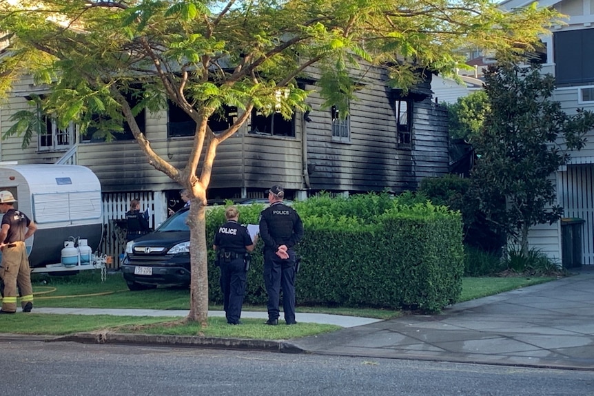 Two police officers stand by a gutted house, with other emergency officers also nearby.