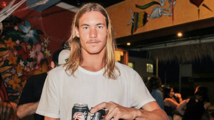 a young man with long hair and a moustache carrying tins of beers looking at the camera