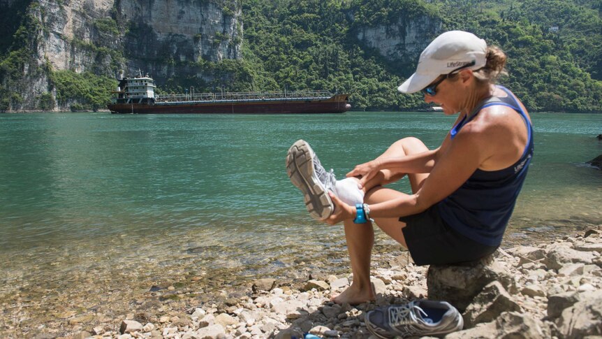 Woman sits on the rocky bank of a lake and removes running shoe. There lake is surrounded by a cliff.