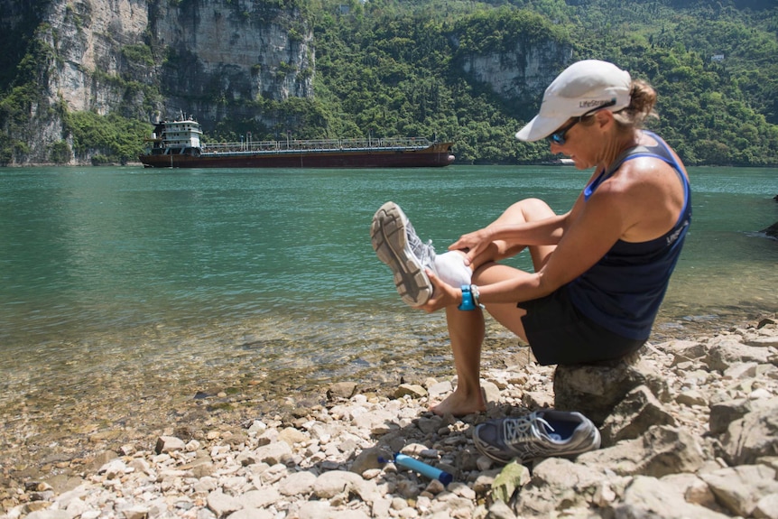 Woman sits on the rocky bank of a lake and removes running shoe. There lake is surrounded by a cliff.