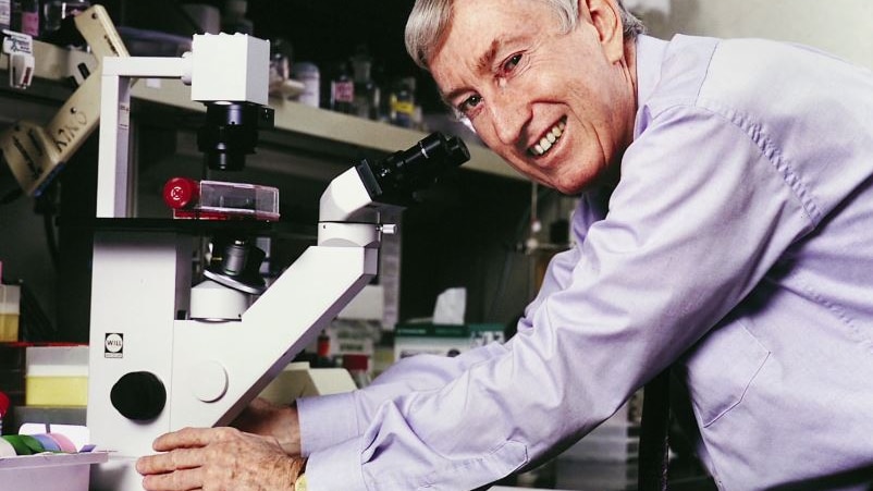 Peter Doherty smiles at the camera while leaning over a microscope.