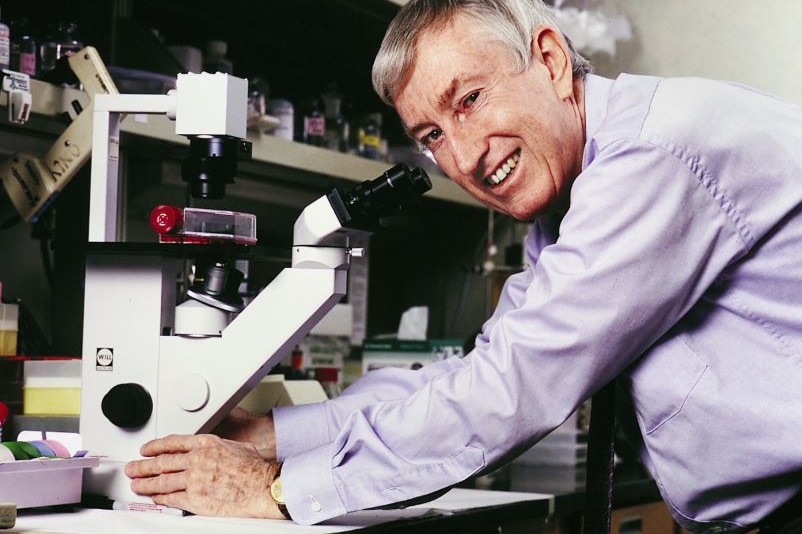 Peter Doherty smiles at the camera while leaning over a microscope.