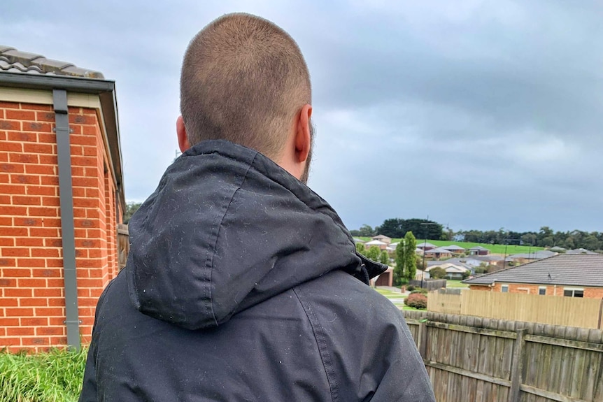 The back of a young man wearing a black coat standing outside a house.