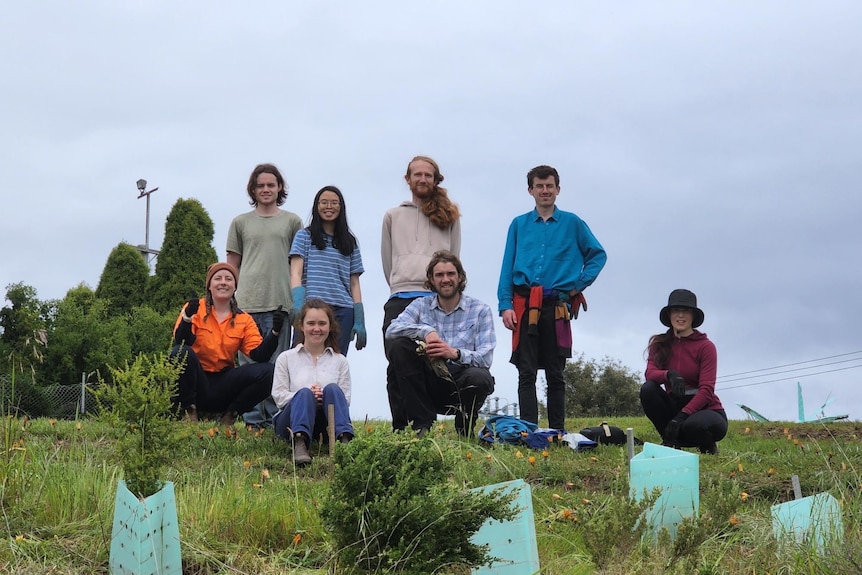 A group of eight people smile and look down to a camera as they stand in a field on an overcast day.