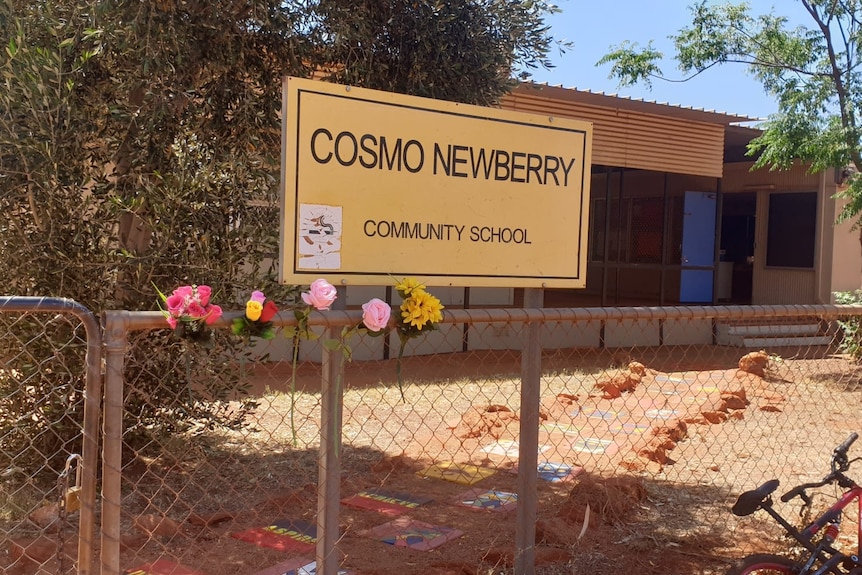 Flowers sit in a chain-link fence, in front of a sign reading COSMO NEWBERRY COMMUNITY SCHOOL