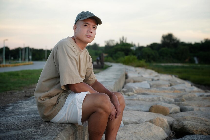 A man wearing a cap sits on a rocky ledge while dressed in a shirt and shorts.