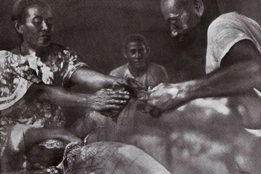 Black and white image of man receiving a pe'a from a master tatooist and woman holding his back. 