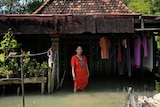 a woman stands outside her flooded home, lifting her dress above knee-high water
