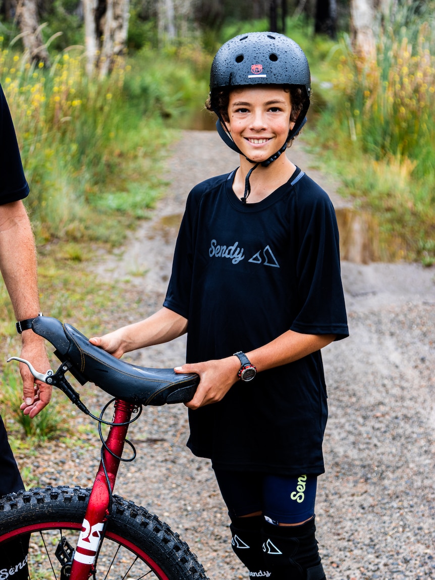 A boy stands in a helmet and cycle gear holding a unicycle on a bush track.