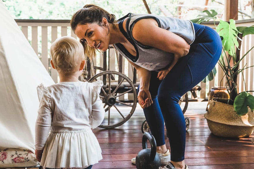 A woman in workout gear standing on a verandah bends down smiling to talk to a toddler