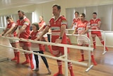 Three rows of footballers of different ages and genders, in uniform, line up at three barres doing ballet poses.