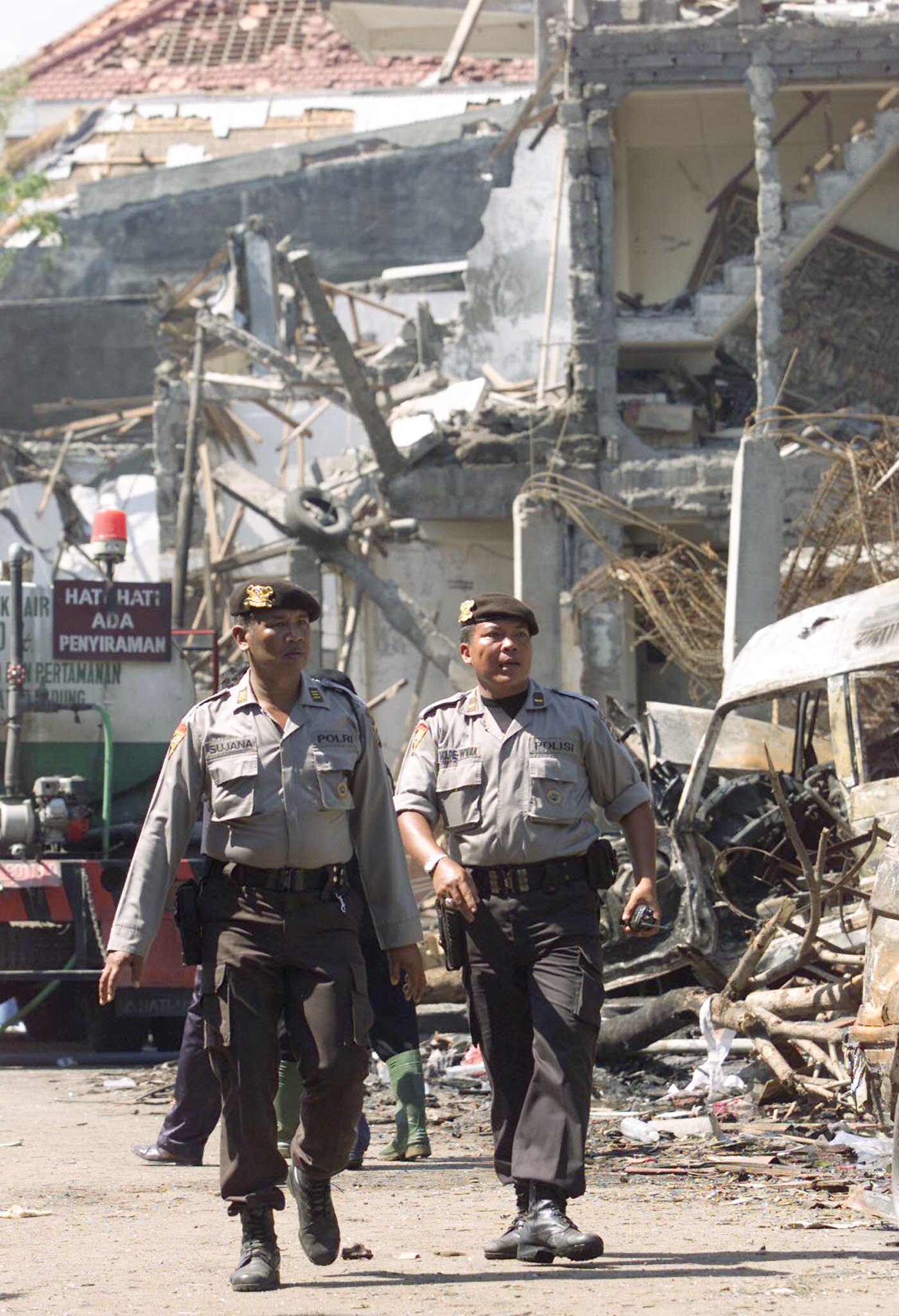 Two men dressed in police uniform and wearing hats walk past a destroyed building.