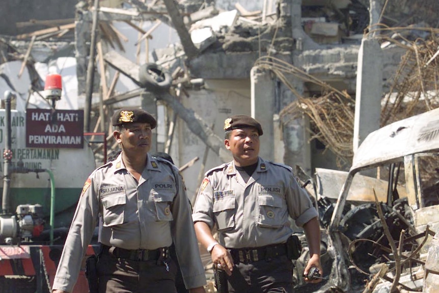 Two men dressed in police uniform and wearing hats walk past a destroyed building.