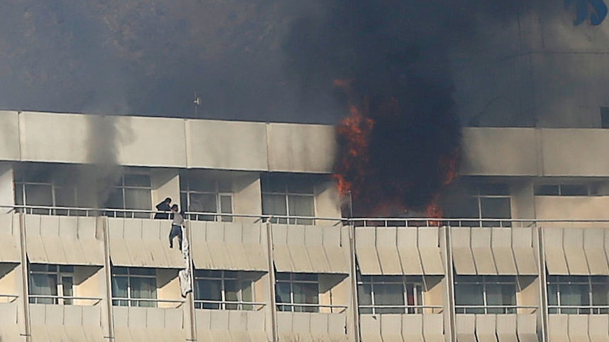 Black smoke and flames spew out of a top floor room while a man hangs off a nearby balcony clinging to bedsheets.