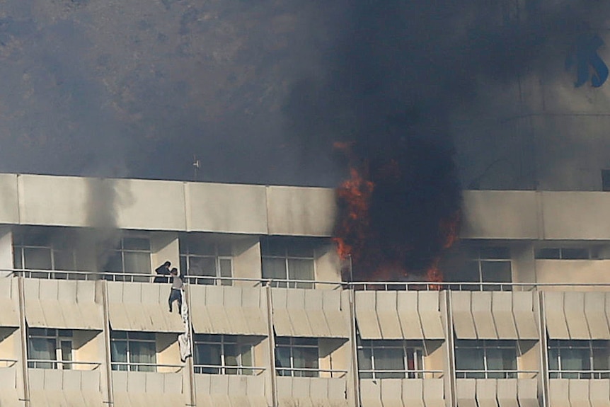 Black smoke and flames spew out of a top floor room while a man hangs off a nearby balcony clinging to bedsheets.