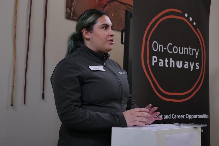 A young woman with black hair dyed with strips of green gives a speech at a podium in front of a sign saying on-country pathways